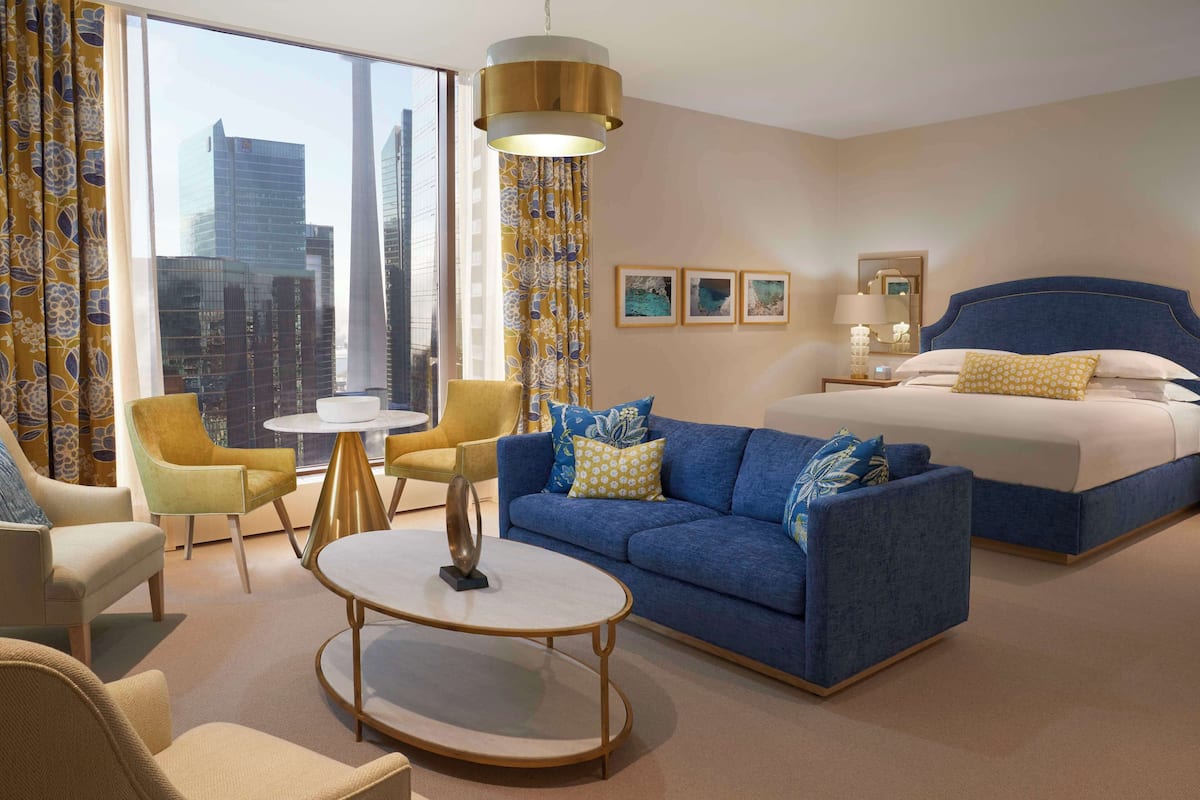 A hotel room with a large bed, a blue sofa, a round coffee table, and a city view through large windows.