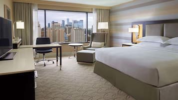 A hotel room with a large bed, a desk with a computer, a chair, a small table, and a view of the city skyline through large windows.