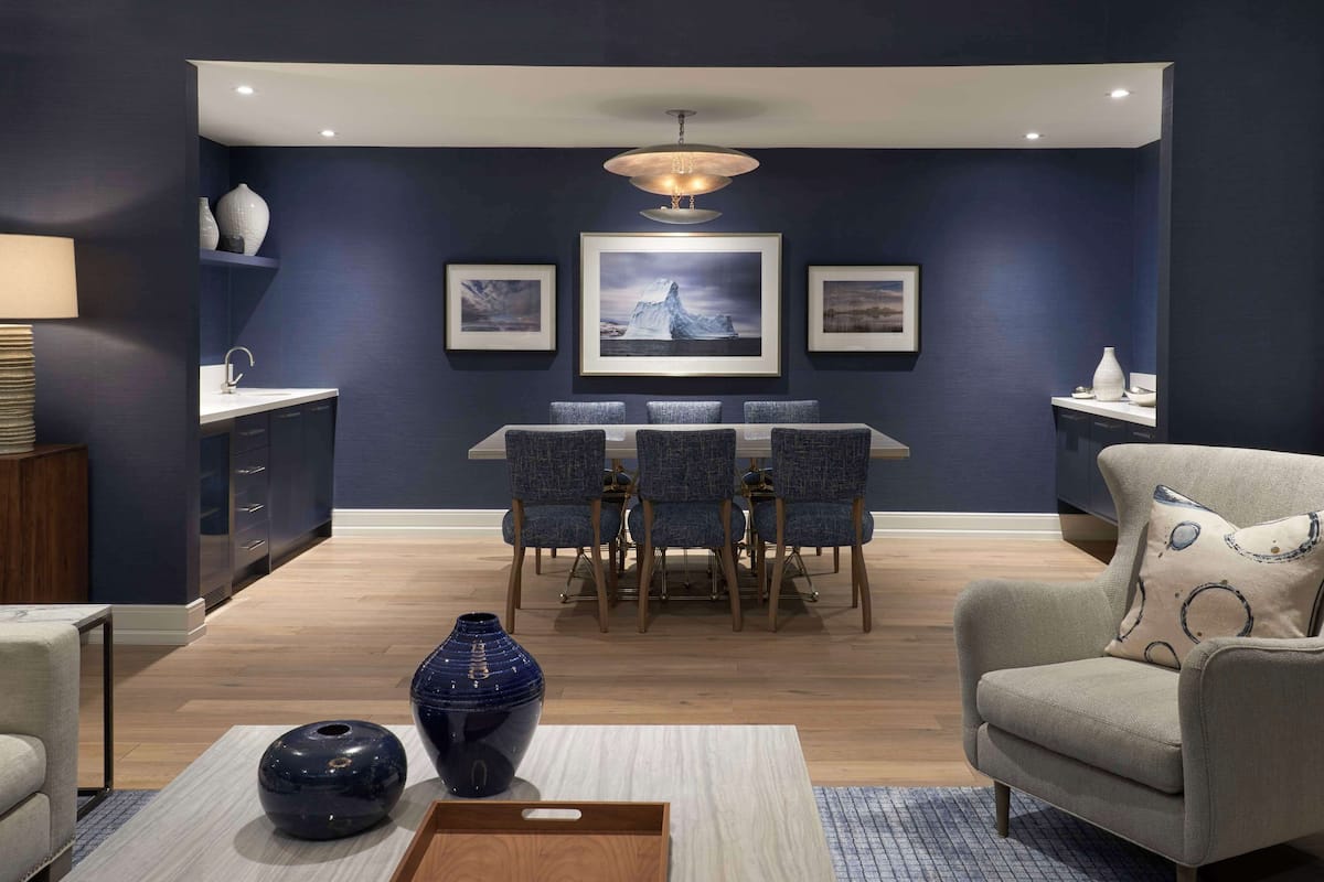 A modern living room with a dining area, featuring a dark blue wall, a chandelier, framed artwork, and a wooden coffee table.