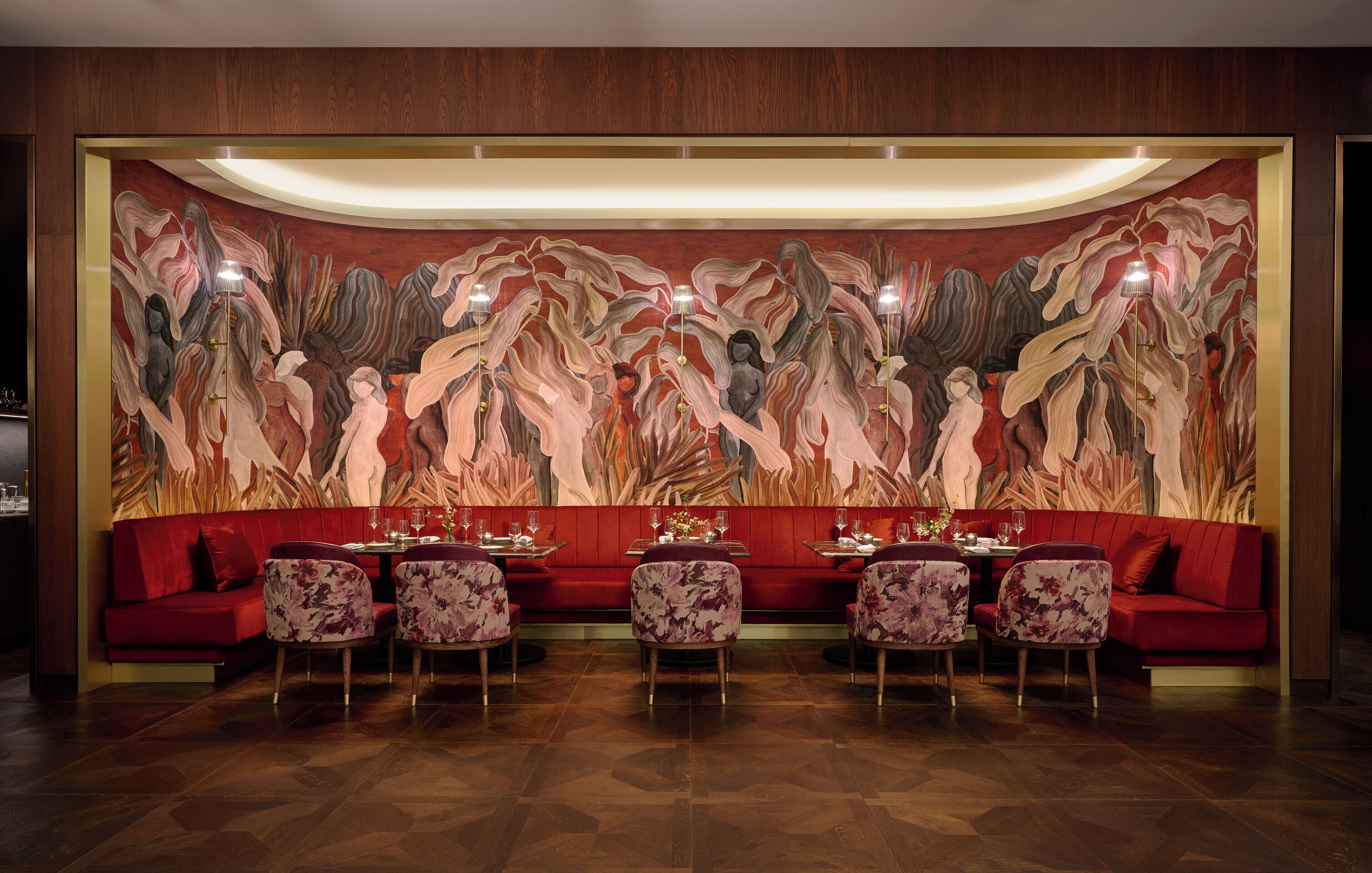 A restaurant with a red sofa and floral chairs, a large abstract mural on the wall, and a wooden floor.
