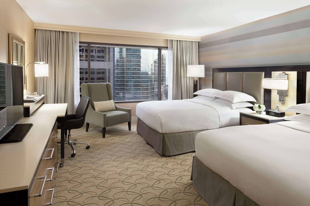 A hotel room with a bed, a desk with a computer, a chair, a nightstand, and a large window with a city view.
