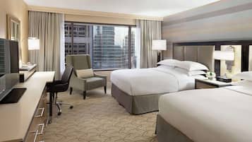 A hotel room with a bed, a desk with a computer, a chair, a nightstand, and a large window with a city view.