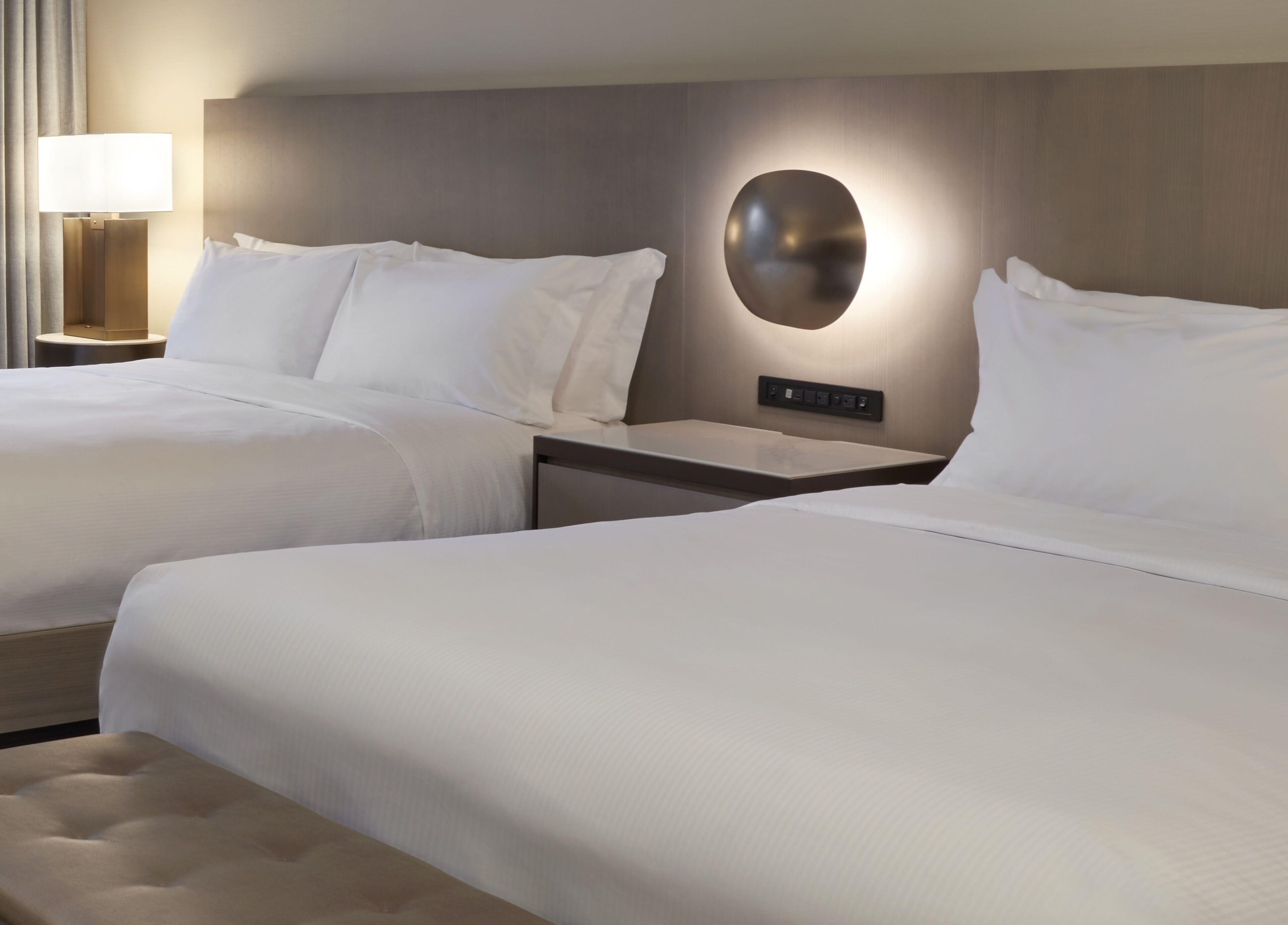 A hotel room with two beds, a bedside table with a lamp, and a wall-mounted control panel.