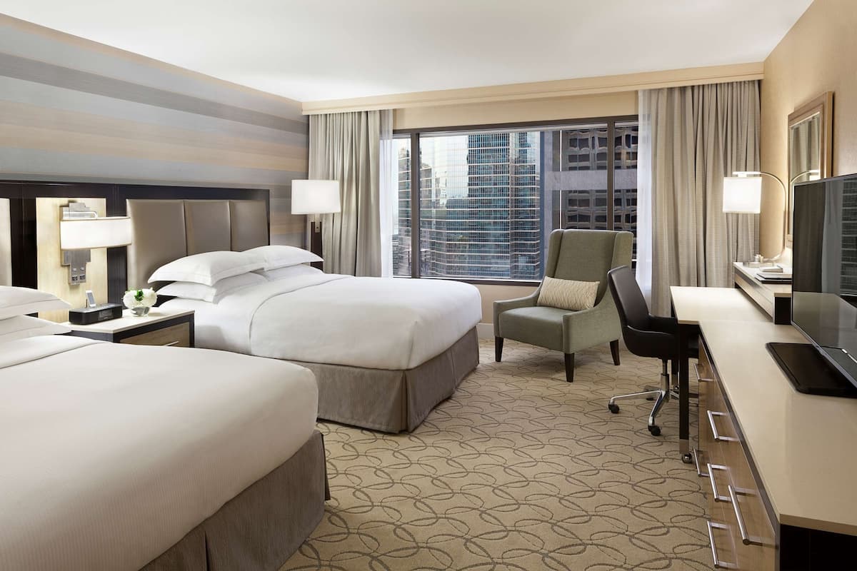 A hotel room with two beds, a desk, a chair, and a large window with a city view.