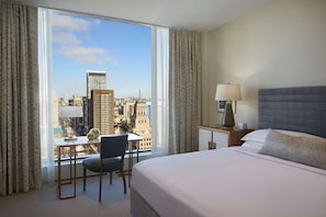 A hotel room with a large window offering a city view, a bed, a desk with a chair, and a nightstand with a lamp.