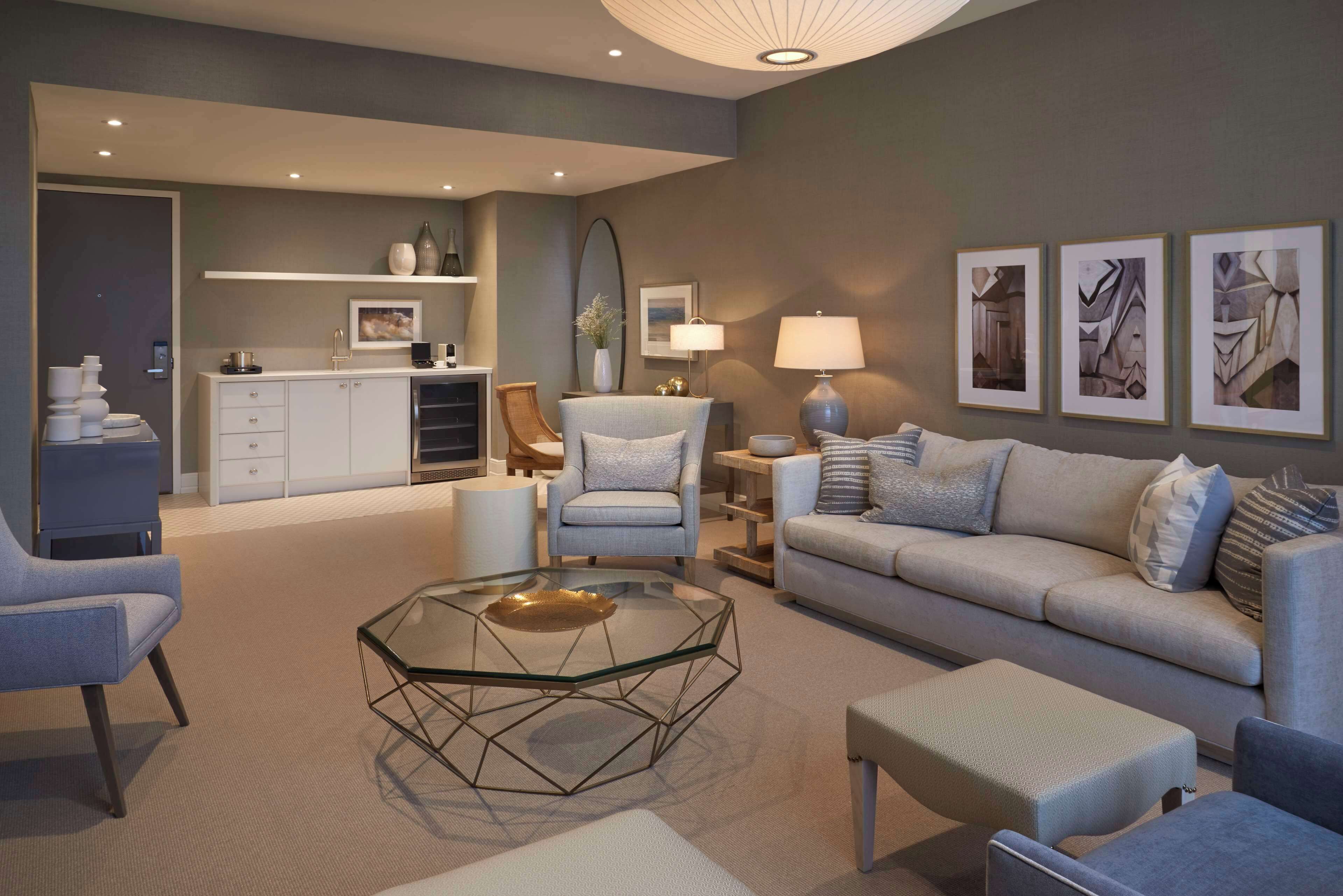 A modern living room with a sofa, armchairs, a glass coffee table, and a dining area with a kitchen in the background.