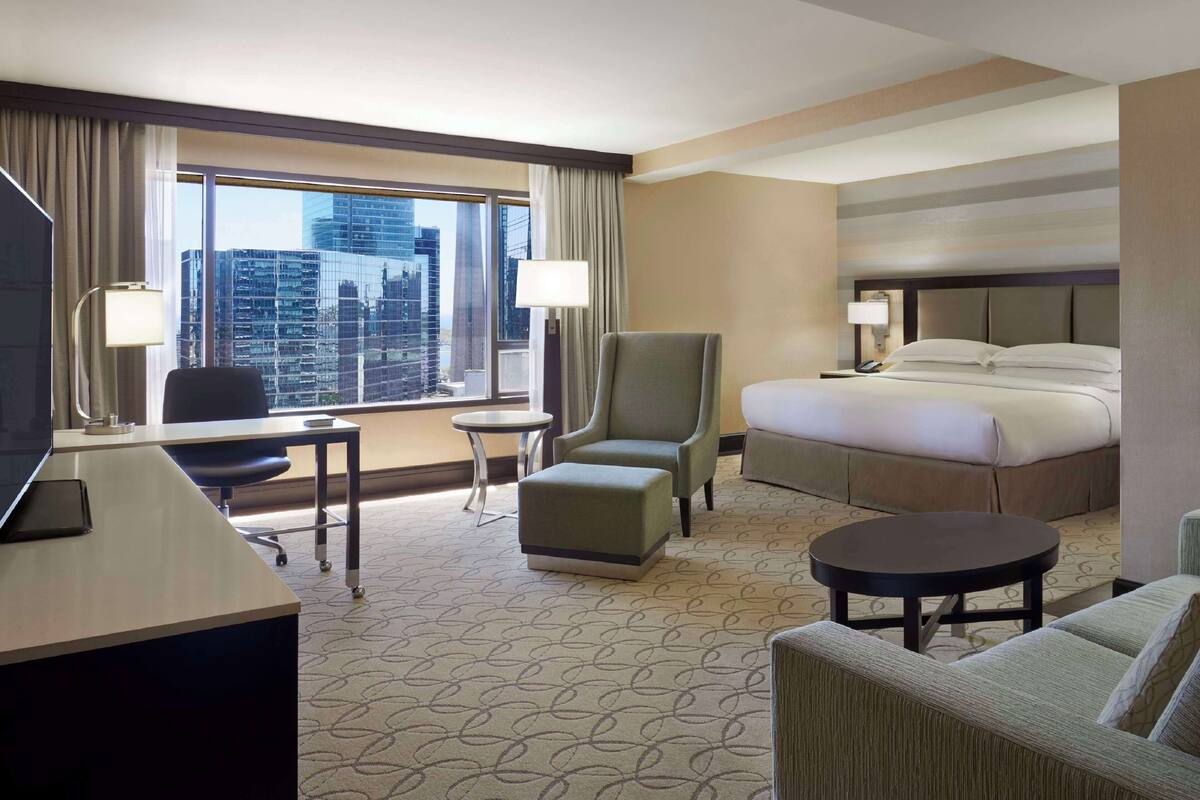 A hotel room with a large bed, a desk with a chair, a small table, a sofa, and a large window offering a city view.