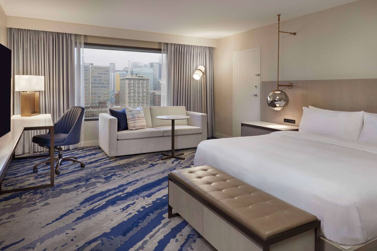 A hotel room with a large bed, a desk, a chair, a sofa, and a view of the city through the window.