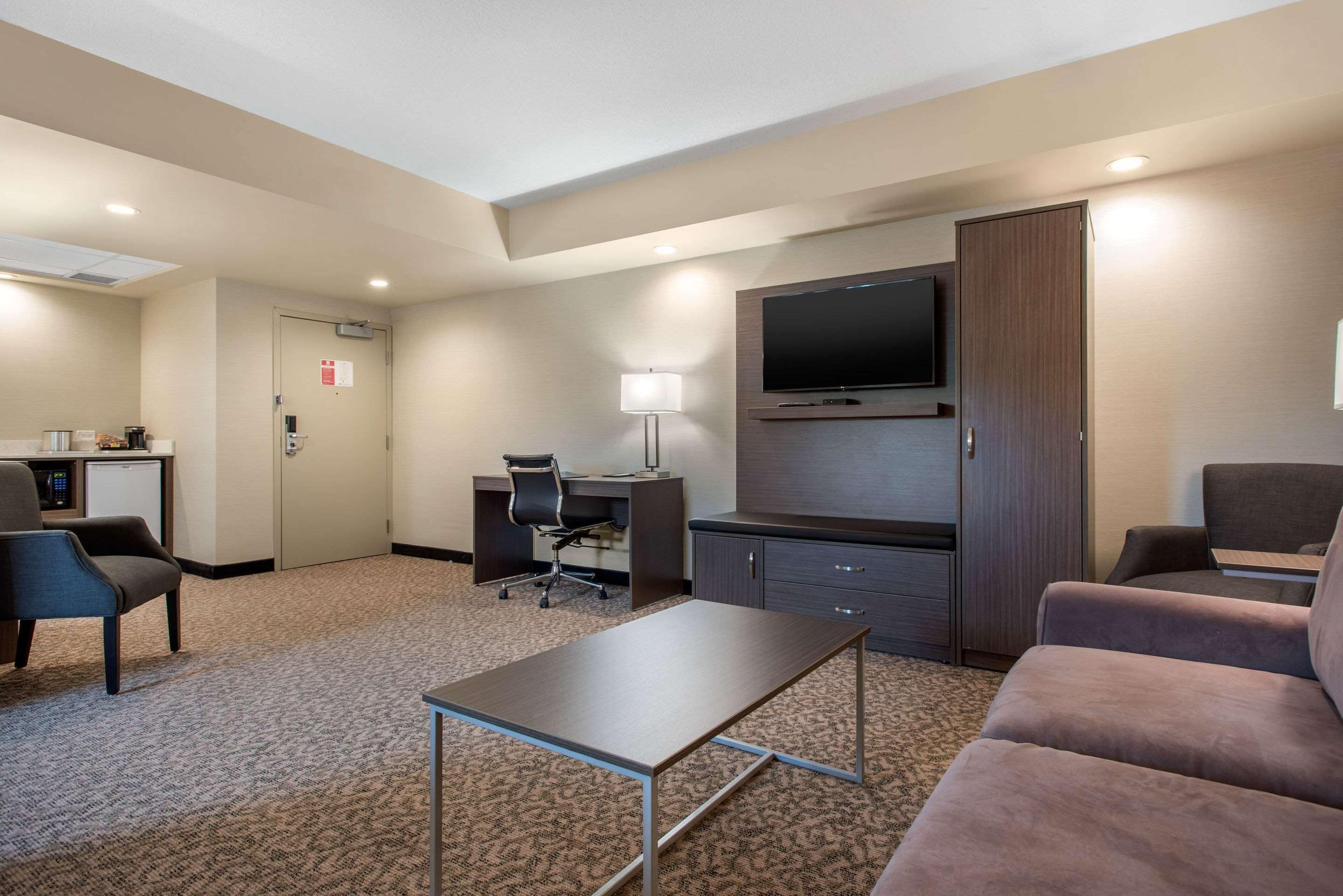 A hotel room with a flat-screen TV mounted above a desk, a sofa, a small table, and a carpeted floor.