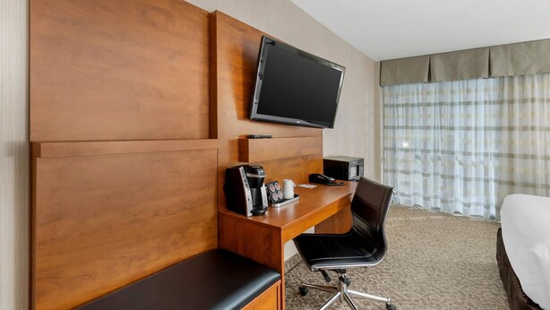 Habitación de hotel con cabecera de madera, televisor de pantalla plana sobre un escritorio con cafetera, una silla y cama con edredón blanco.