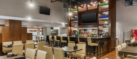 A restaurant with multiple tables and chairs, a bar area with shelves of bottles, and a flat-screen TV mounted on the wall.