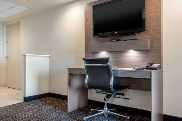 A hotel room with a desk, chair, flat-screen TV, and a small refrigerator.