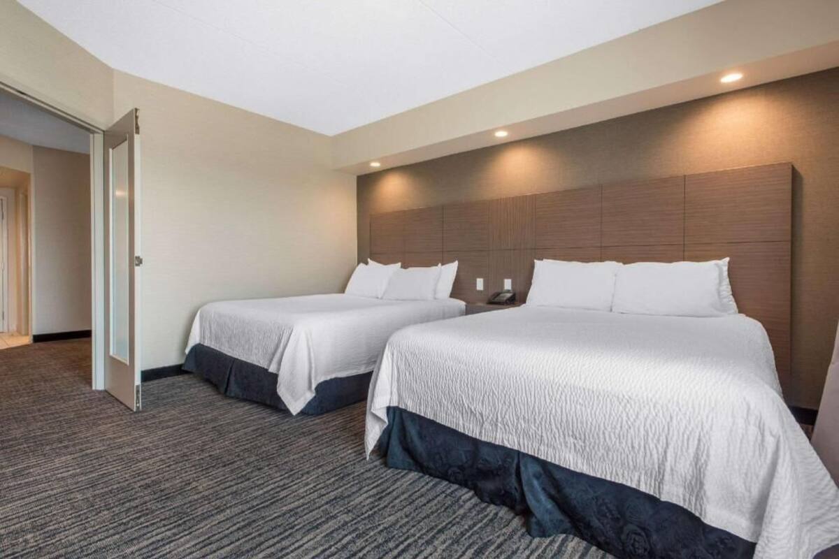 A hotel room with two beds, a wooden headboard, a carpeted floor, and a door leading to another room.