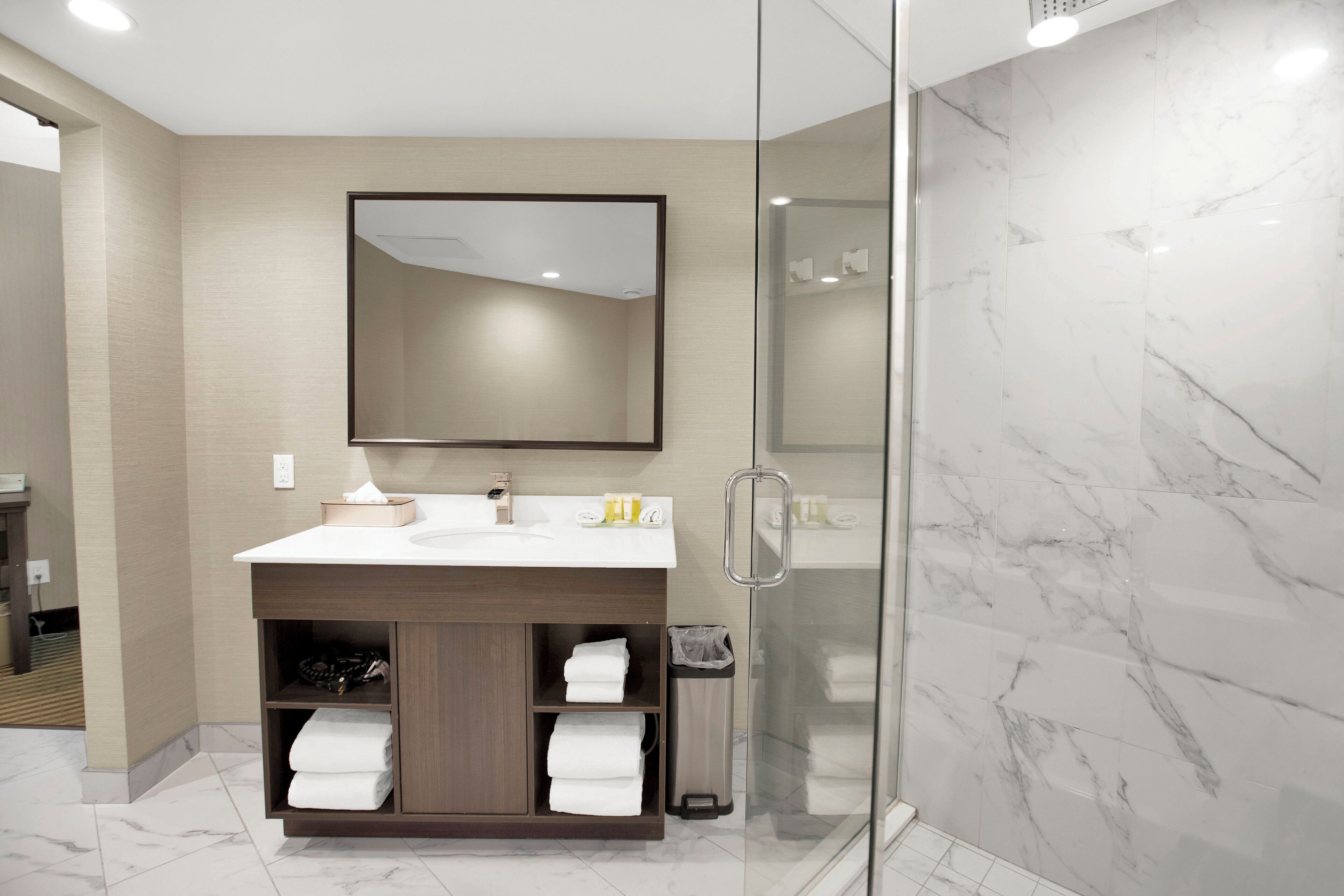 Une salle de bain moderne avec une douche en marbre, une douche à l’italienne avec des parois en verre, un meuble-lavabo en bois avec un miroir et des serviettes soigneusement pliées.
