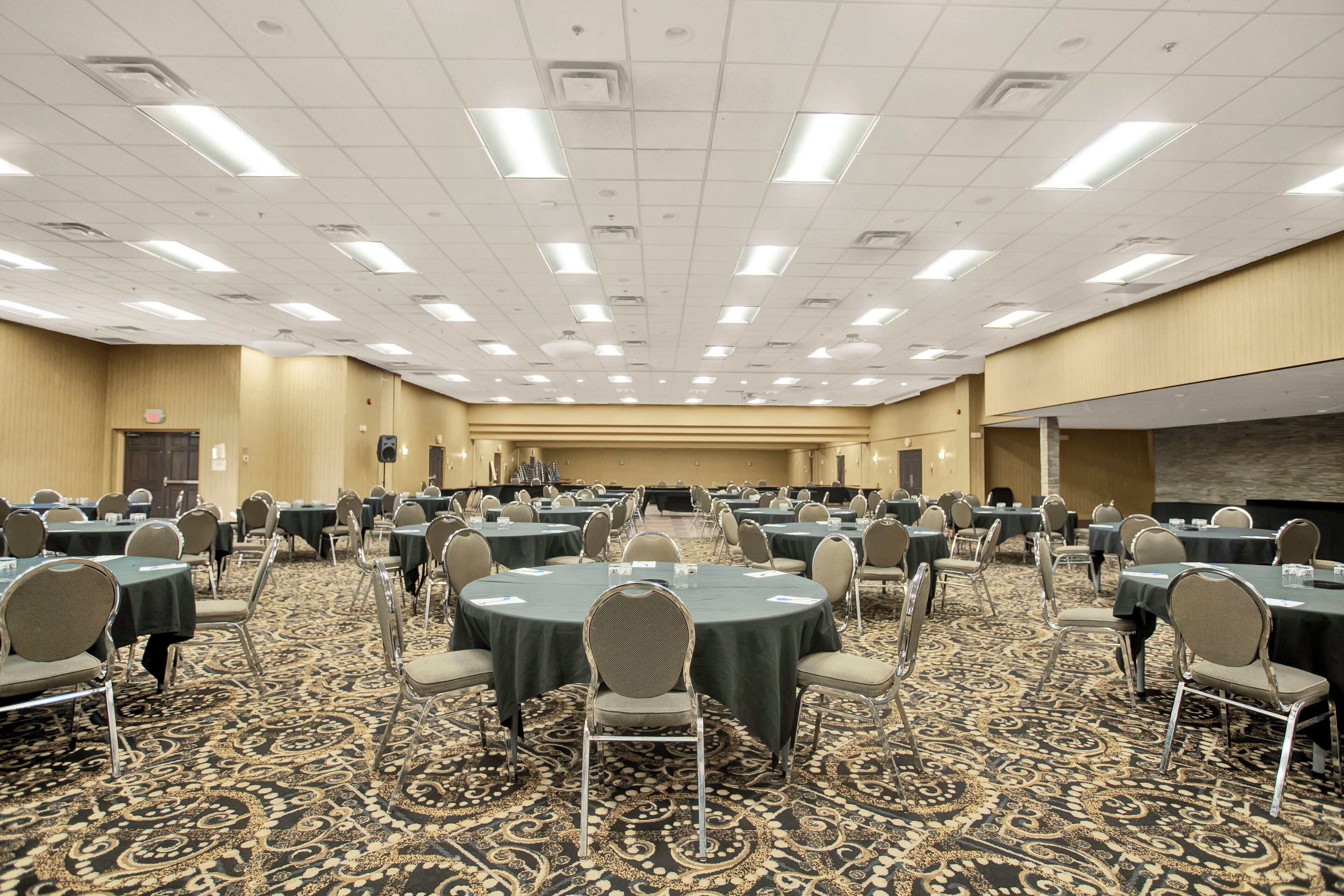 Een ruime congreszaal met ronde tafels en stoelen, een vloerbedekking en een hoog plafond met ingebouwde verlichting.