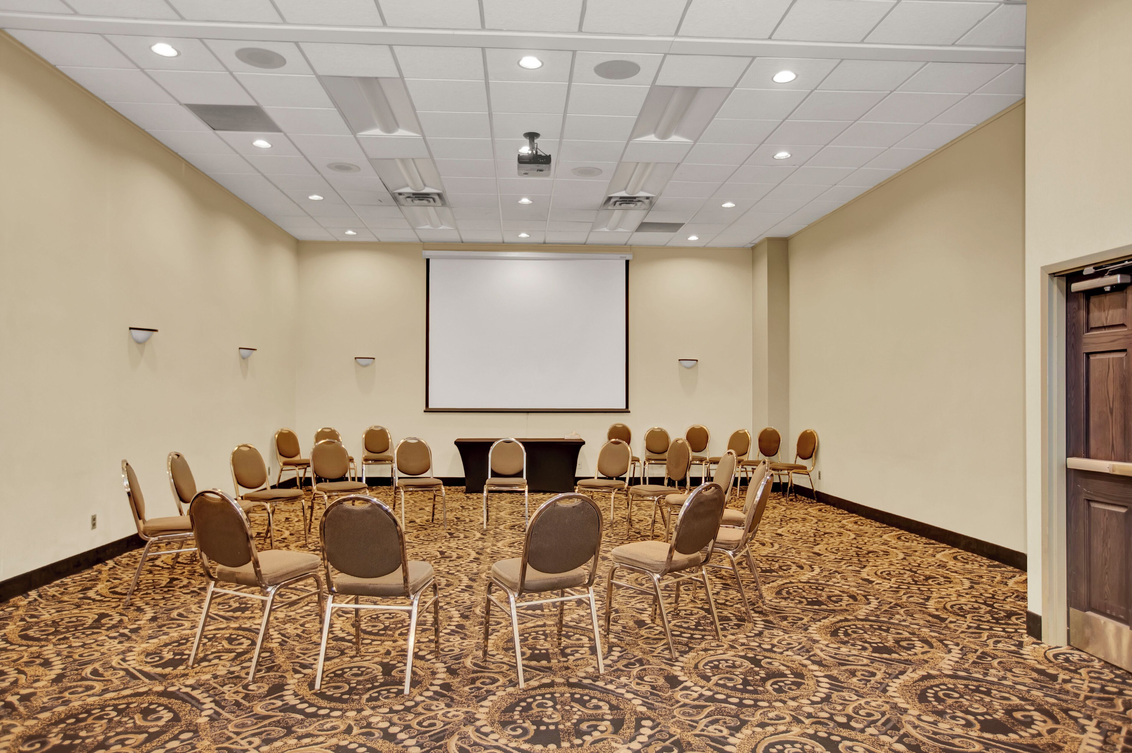 A conference room with a carpeted floor, rows of chairs, a projector screen, and a wooden door.