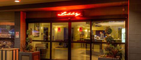 A hotel entrance with a neon sign, glass doors, and potted plants.