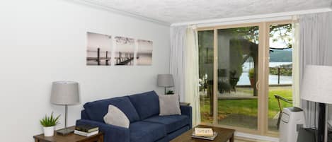 A modern living room with a blue sofa, two side tables with lamps, a wooden coffee table, and a large sliding glass door leading to a balcony with a view of a lake and trees.