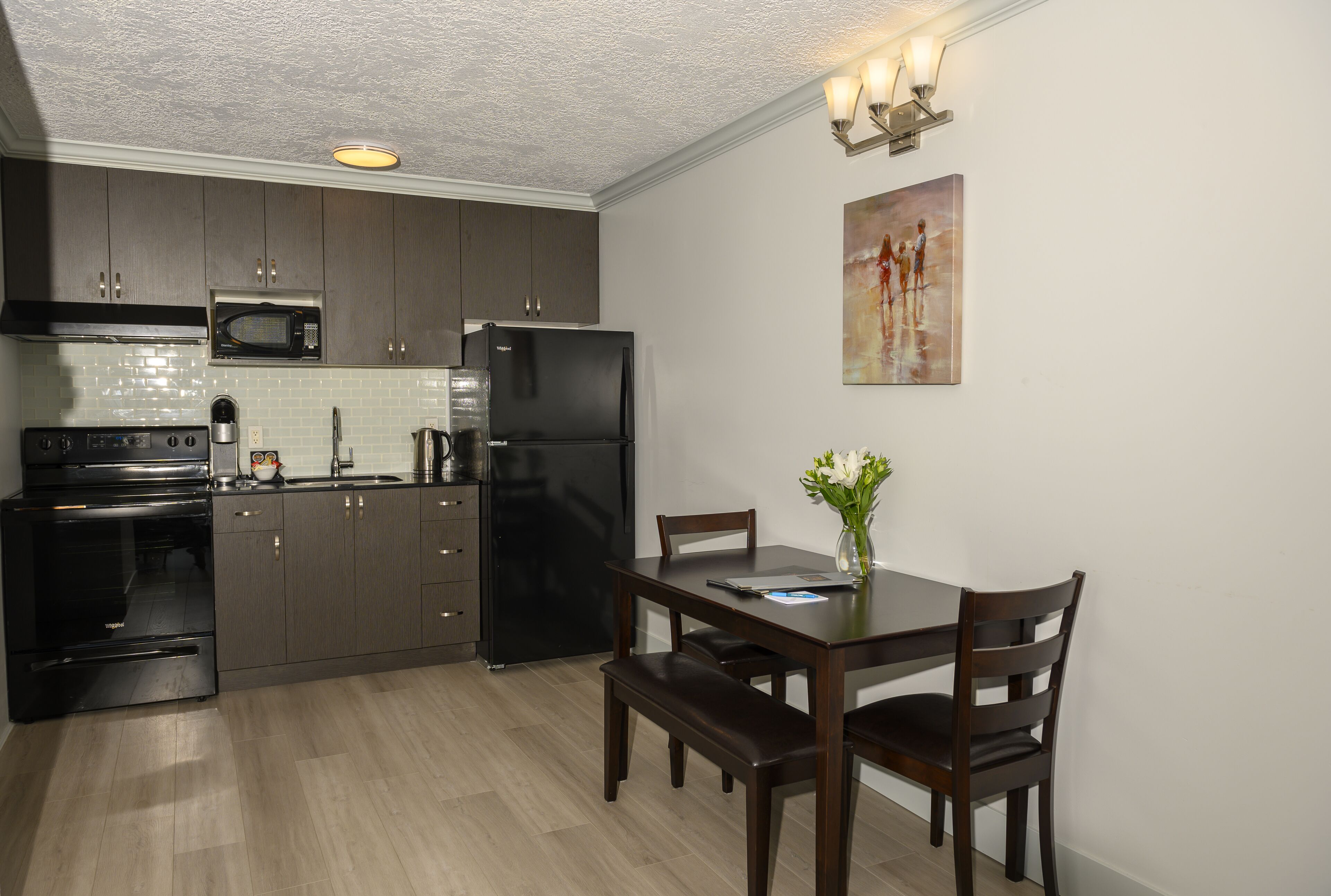 A modern kitchen with dark wood cabinets, a black refrigerator, a microwave, and a stove. There is a dining table with chairs and a vase with flowers.