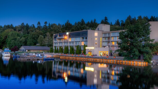 Un hôtel moderne dont l’image se reflète dans l’eau, entouré d’arbres et doté d’un quai où sont amarrés des bateaux.