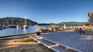 Un port de plaisance avec des bateaux amarrés, une terrasse en bois avec des chaises, et une vue sur des collines en arrière-plan.