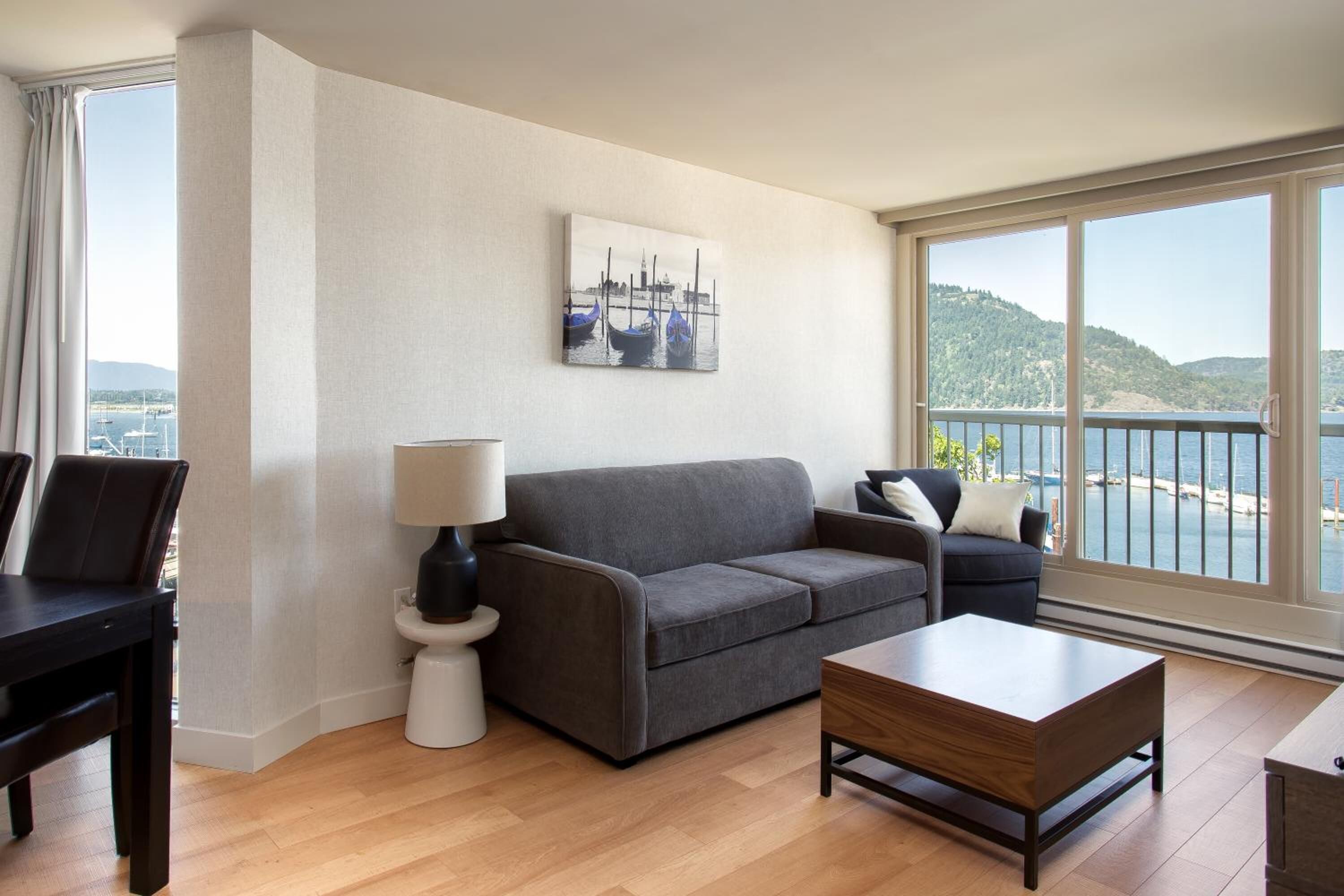 Una sala de estar moderna con un sofá gris, una mesa de centro de madera y vista a un cuerpo de agua a través de amplios ventanales.