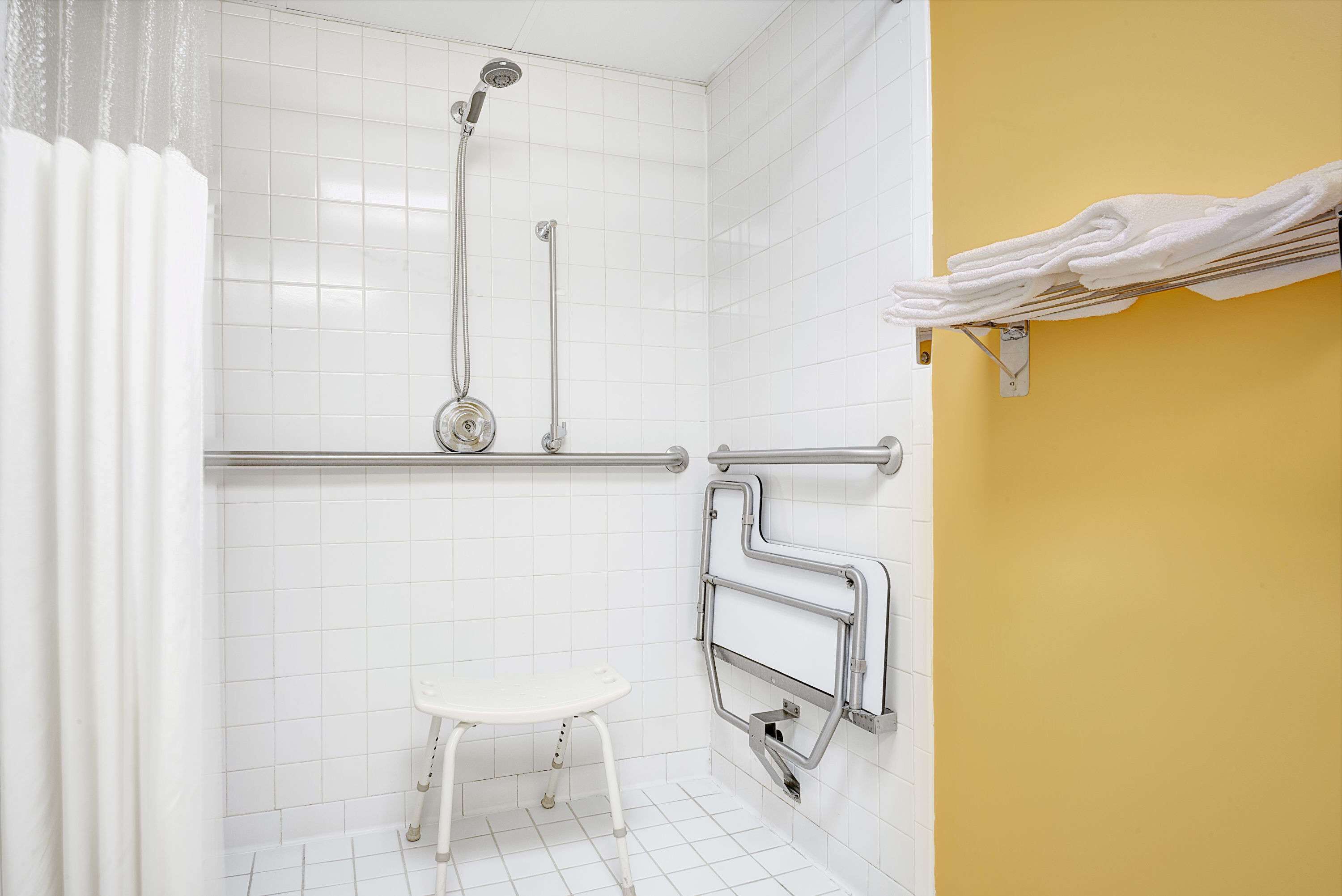 A bathroom with white tiles, a shower with a handheld showerhead, a grab bar, a white stool, and a towel rack.