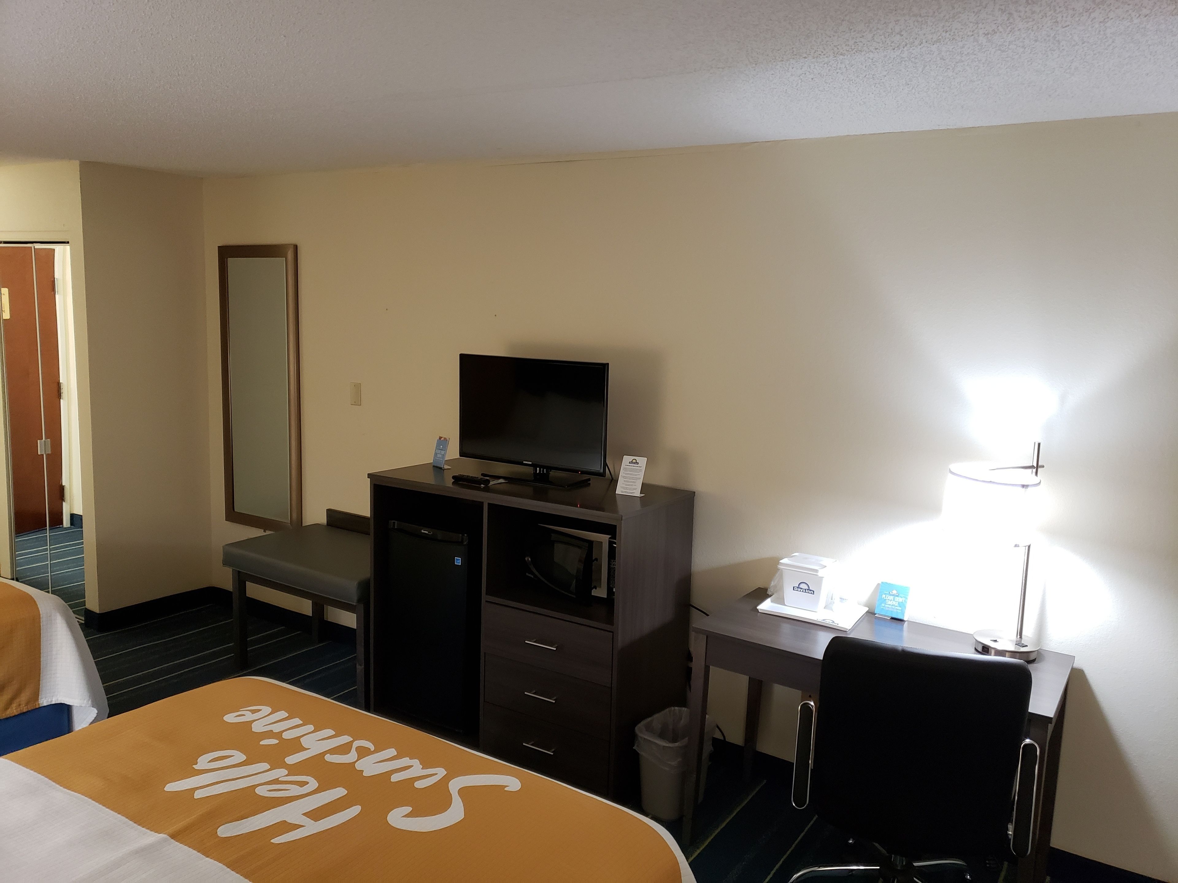 Quarto de hotel com cama, escrivaninha, televisão, lixeira e luminária.