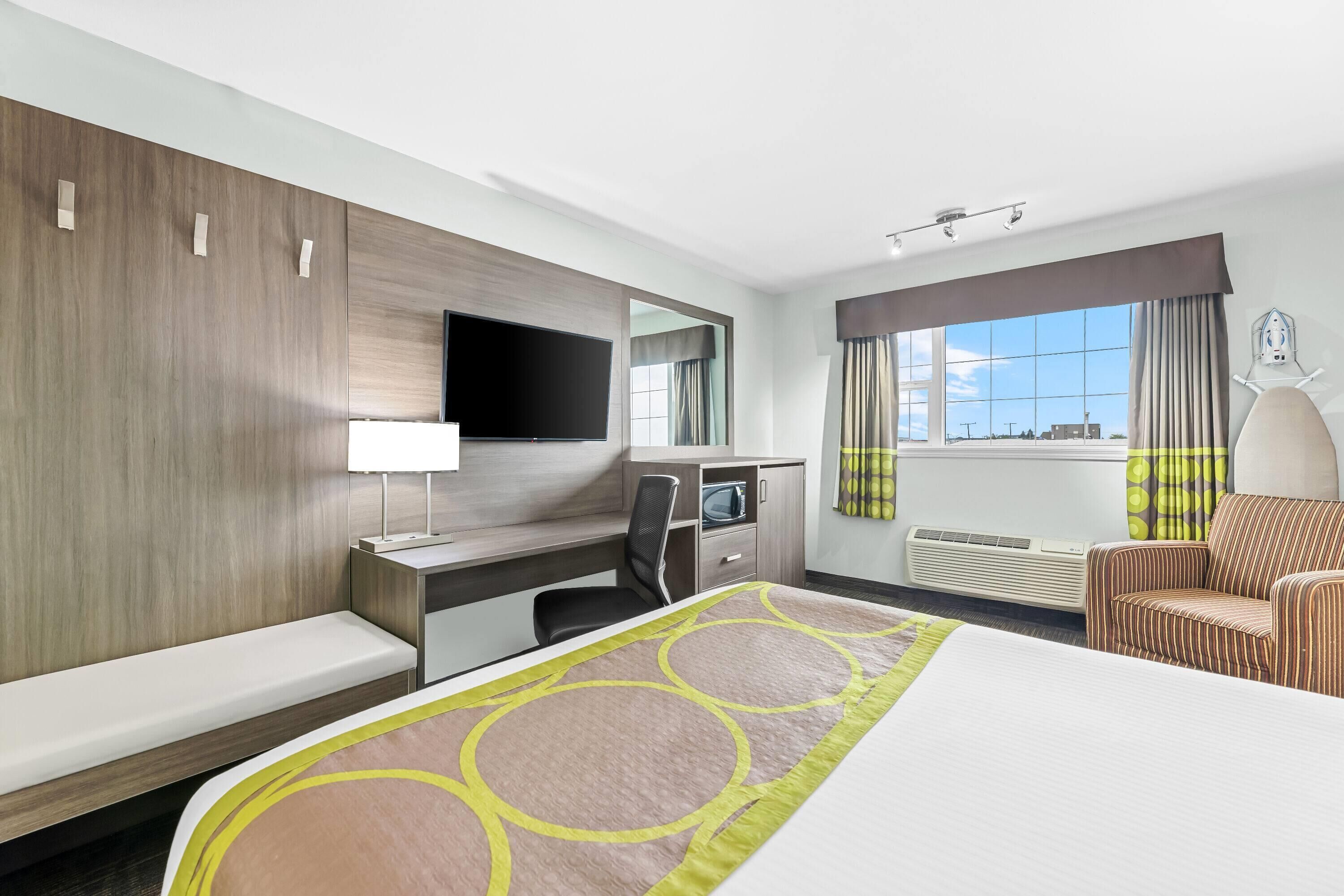A hotel room with a bed, a desk with a flat-screen TV, a chair, a sofa, a window with curtains, and a view of the city.