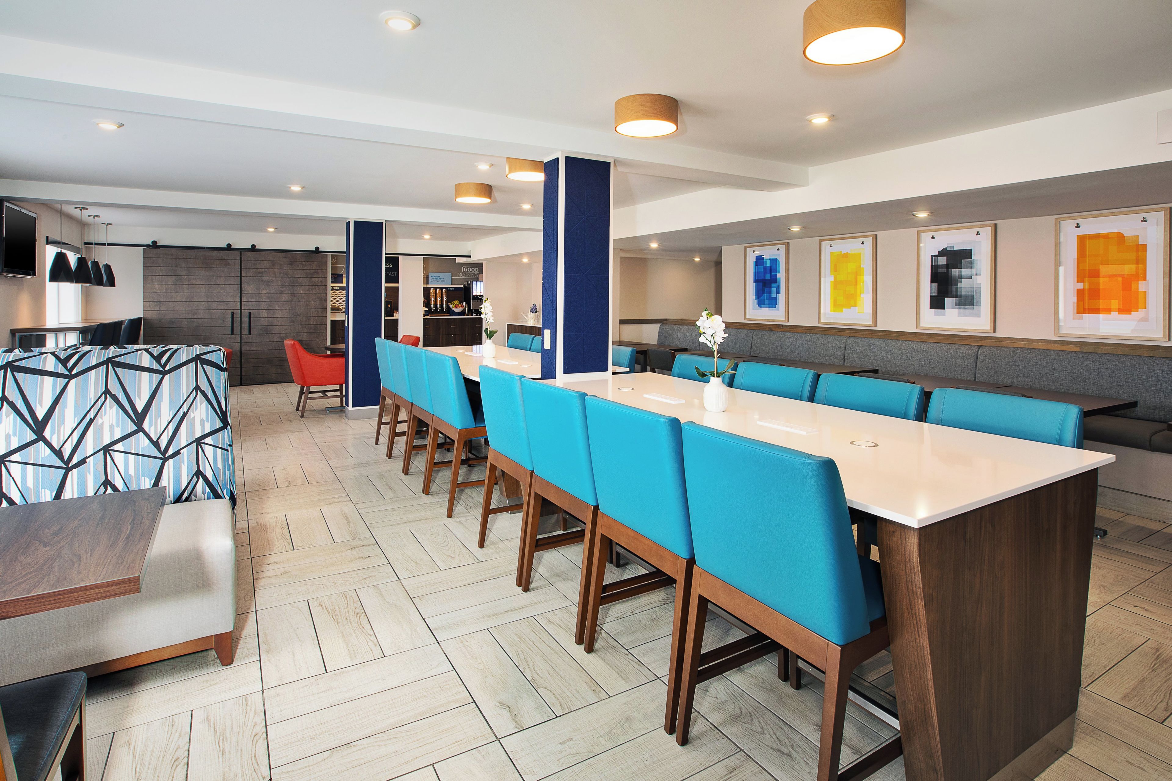 A modern hotel dining area with a long communal table, blue upholstered chairs, and a bar area in the background.