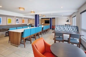 A modern hotel lobby with a long communal table, blue and orange seating, and a large flat-screen TV.