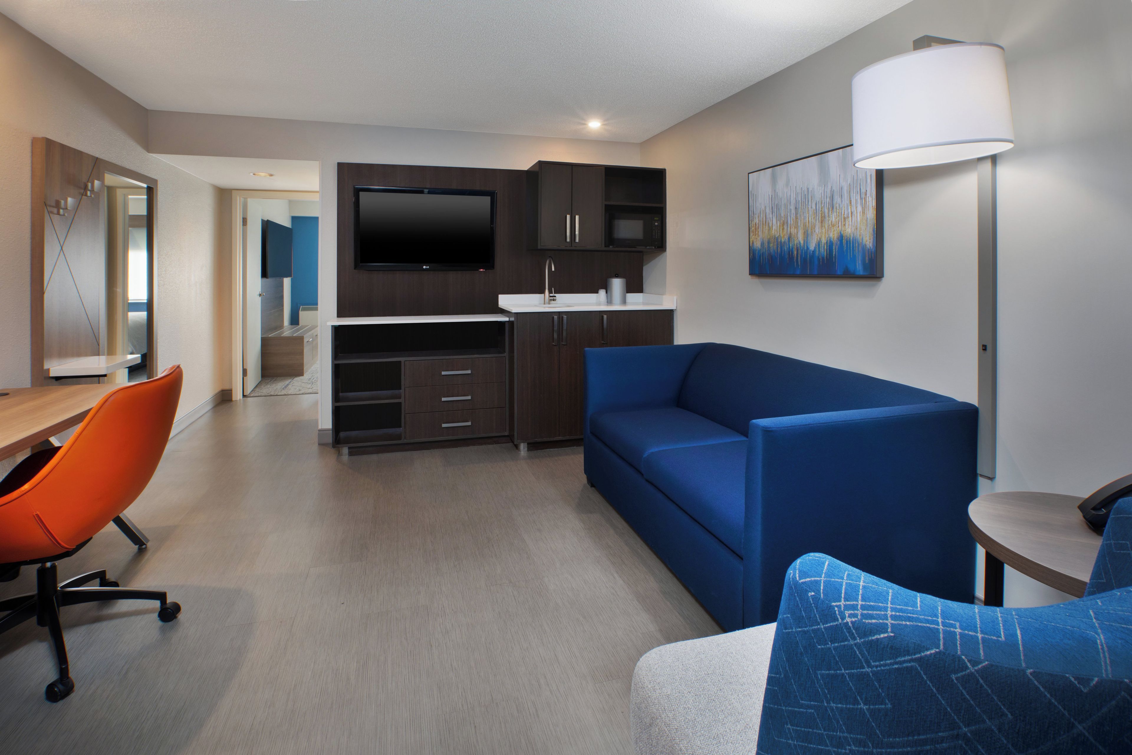 Een moderne hotelkamer met een kitchenette, een flatscreen televisie, een bank en een bureau met een oranje stoel.