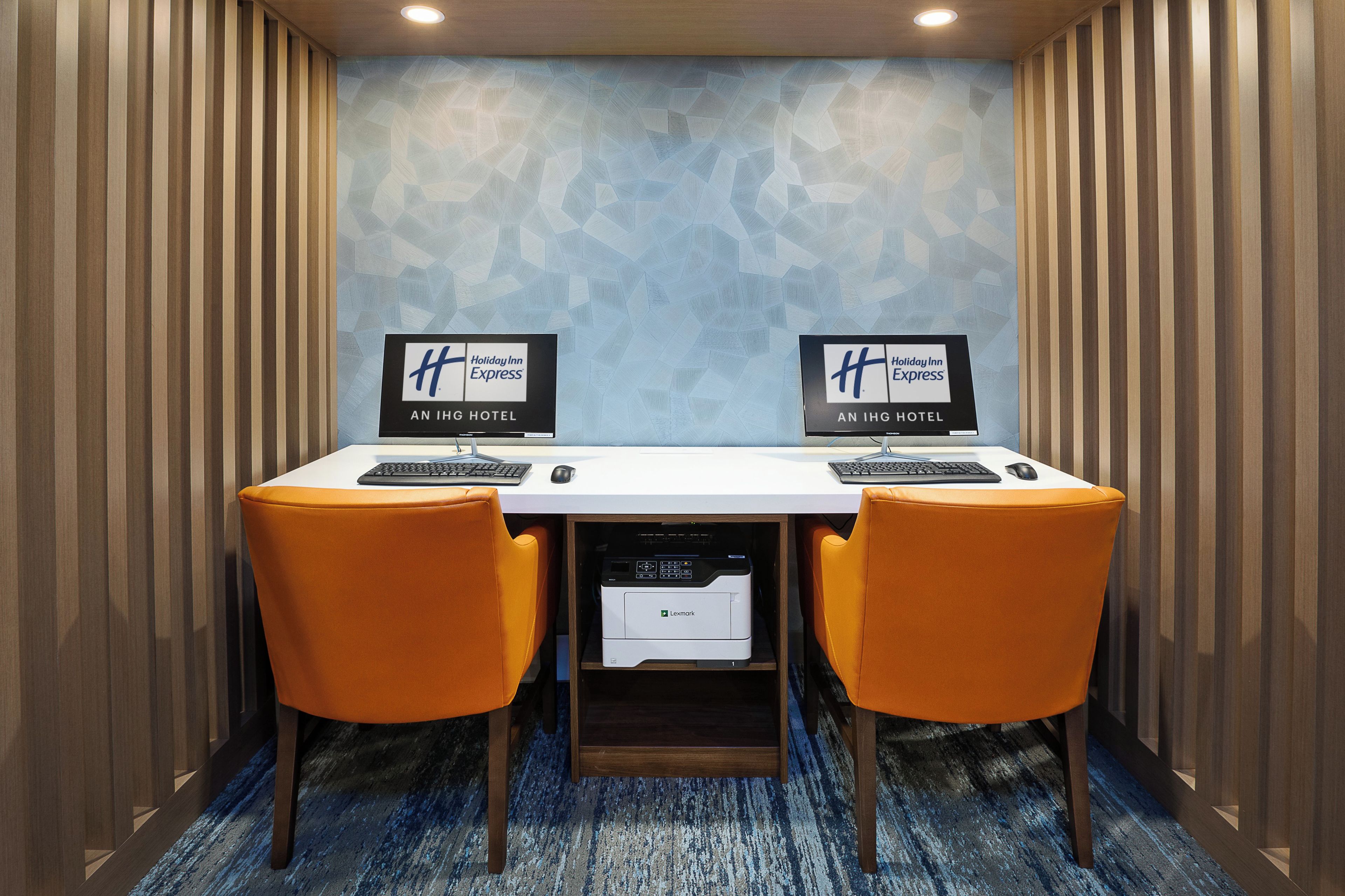Een vergaderruimte met twee bureaus, elk met een Holiday Inn Express bord, twee oranje stoelen en een Lexmark printer.