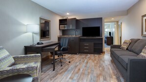 Une chambre d’hôtel moderne avec un plancher en bois, un canapé, un bureau avec une chaise, une télévision et un coin cuisine équipé de placards et d’un four à micro-ondes.
