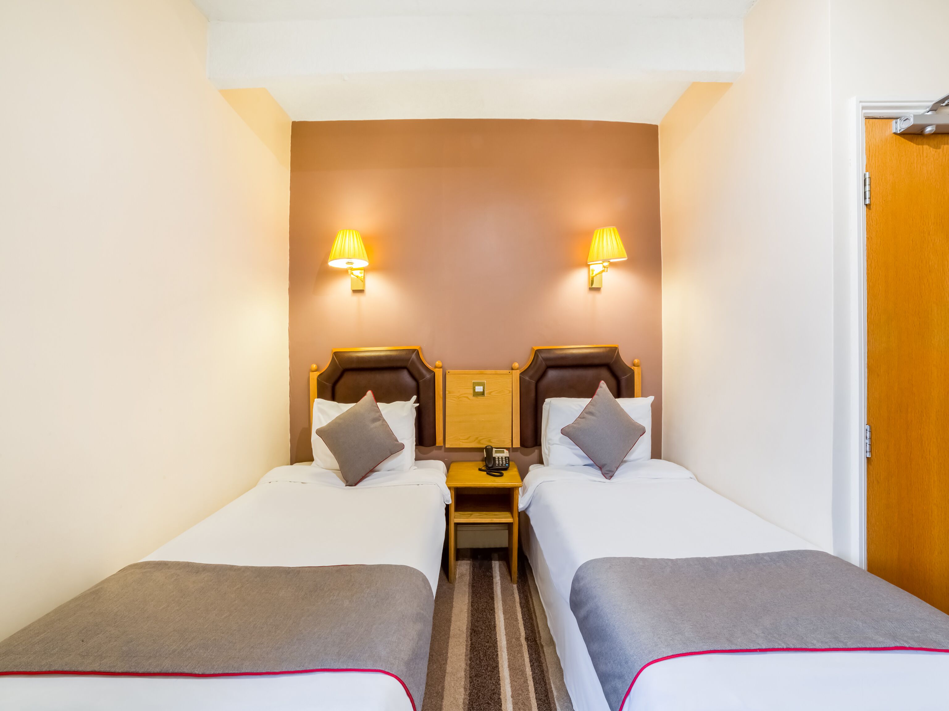 A hotel room with two single beds, each with a pillow and a bedside table. There are two wall-mounted lamps on either side of the beds.