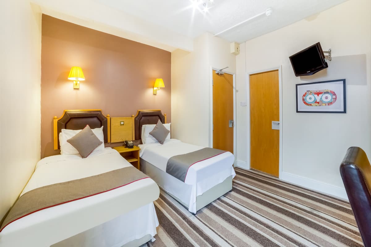 A hotel room with two beds, a bedside table with a lamp, a TV mounted on the wall, and a framed picture.
