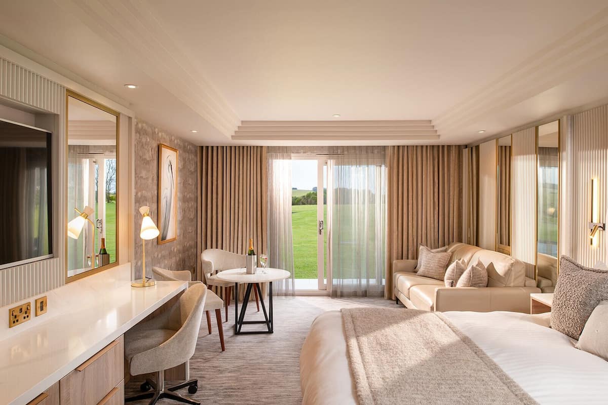 A hotel room with a bed, a desk, a chair, a table with a bottle and glasses, and a large window with curtains.