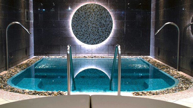 A modern indoor swimming pool with a central circular mosaic, surrounded by a pebble-lined edge and equipped with two water jets.