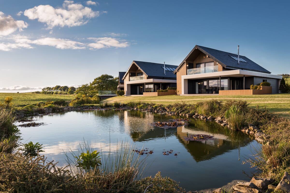 A modern house with a large pond in front, surrounded by greenery and a clear sky.