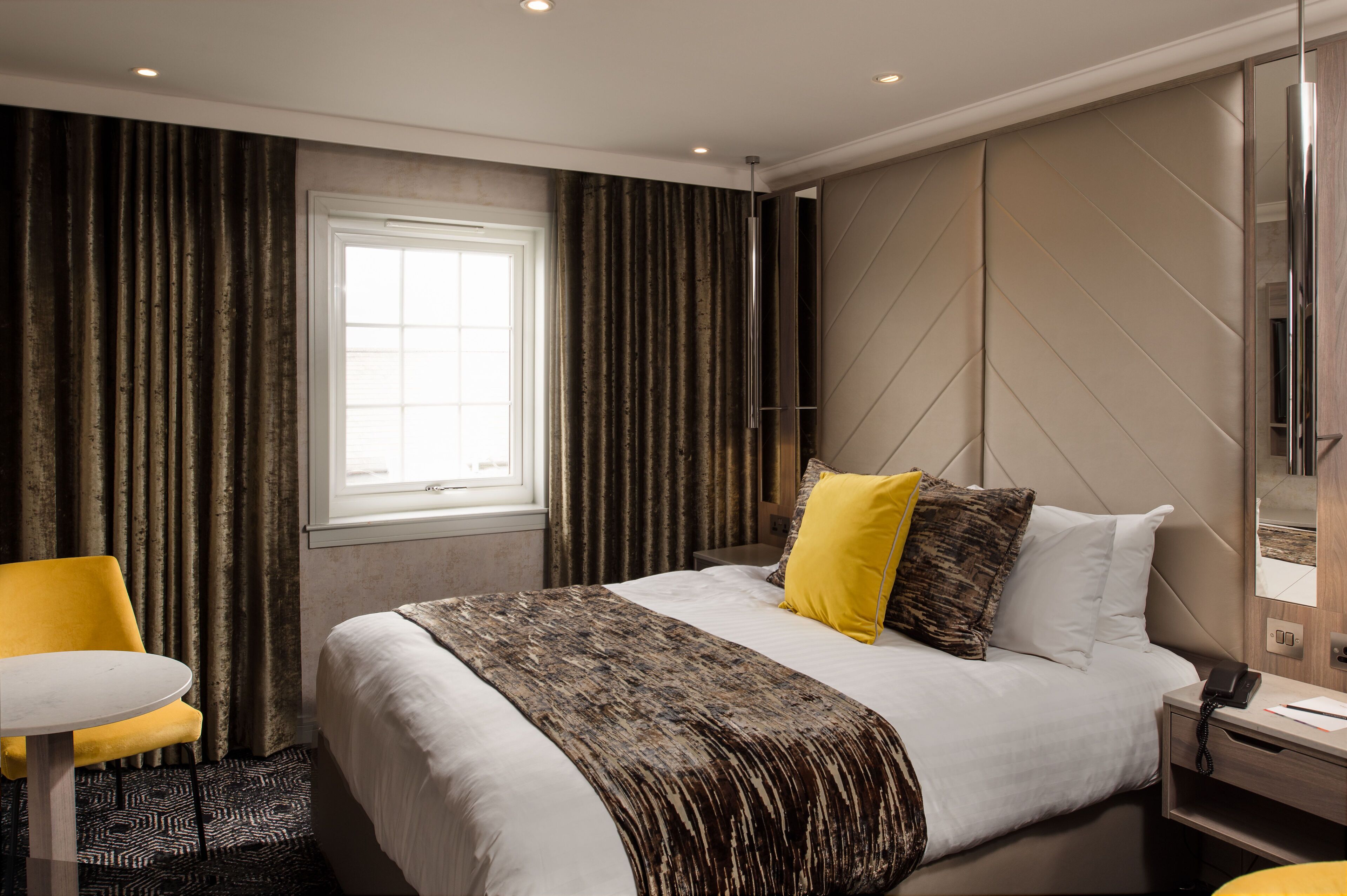 A modern hotel room with a large bed, a bedside table, a chair, and a window with curtains.