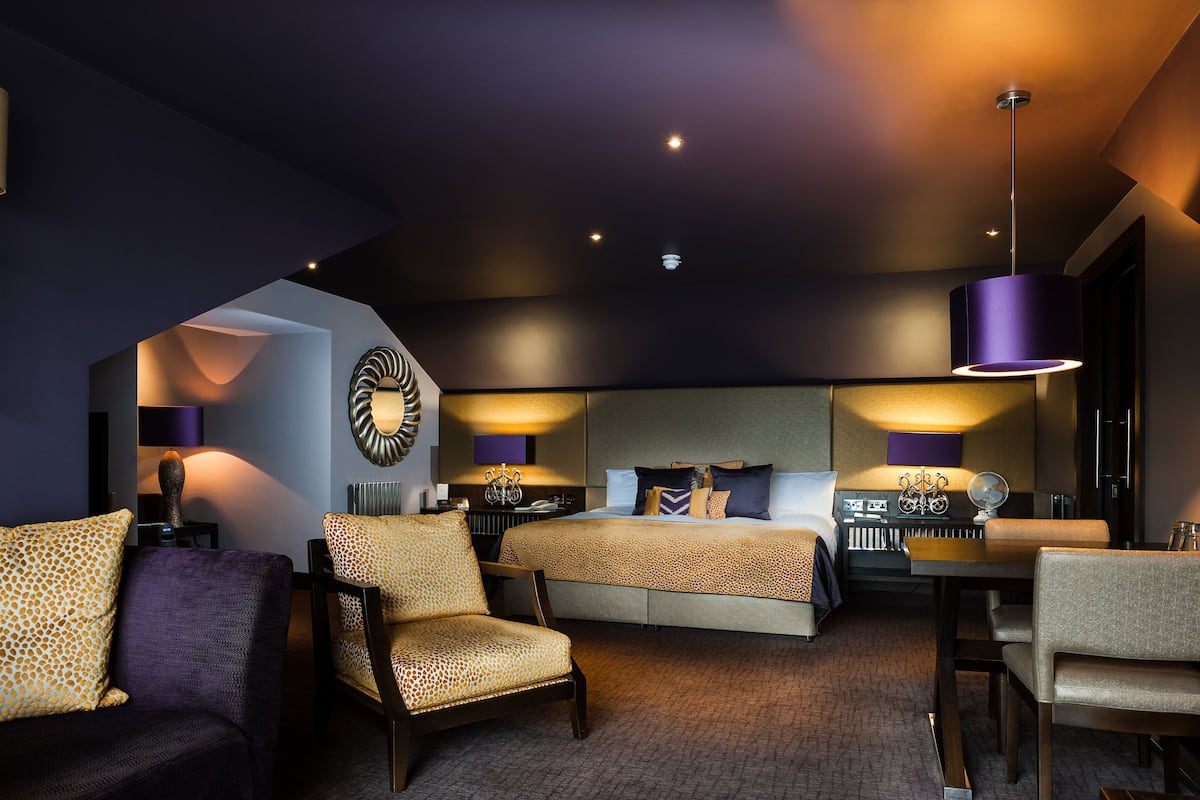 A modern hotel room with a large bed, two armchairs, a desk, and a ceiling light fixture.