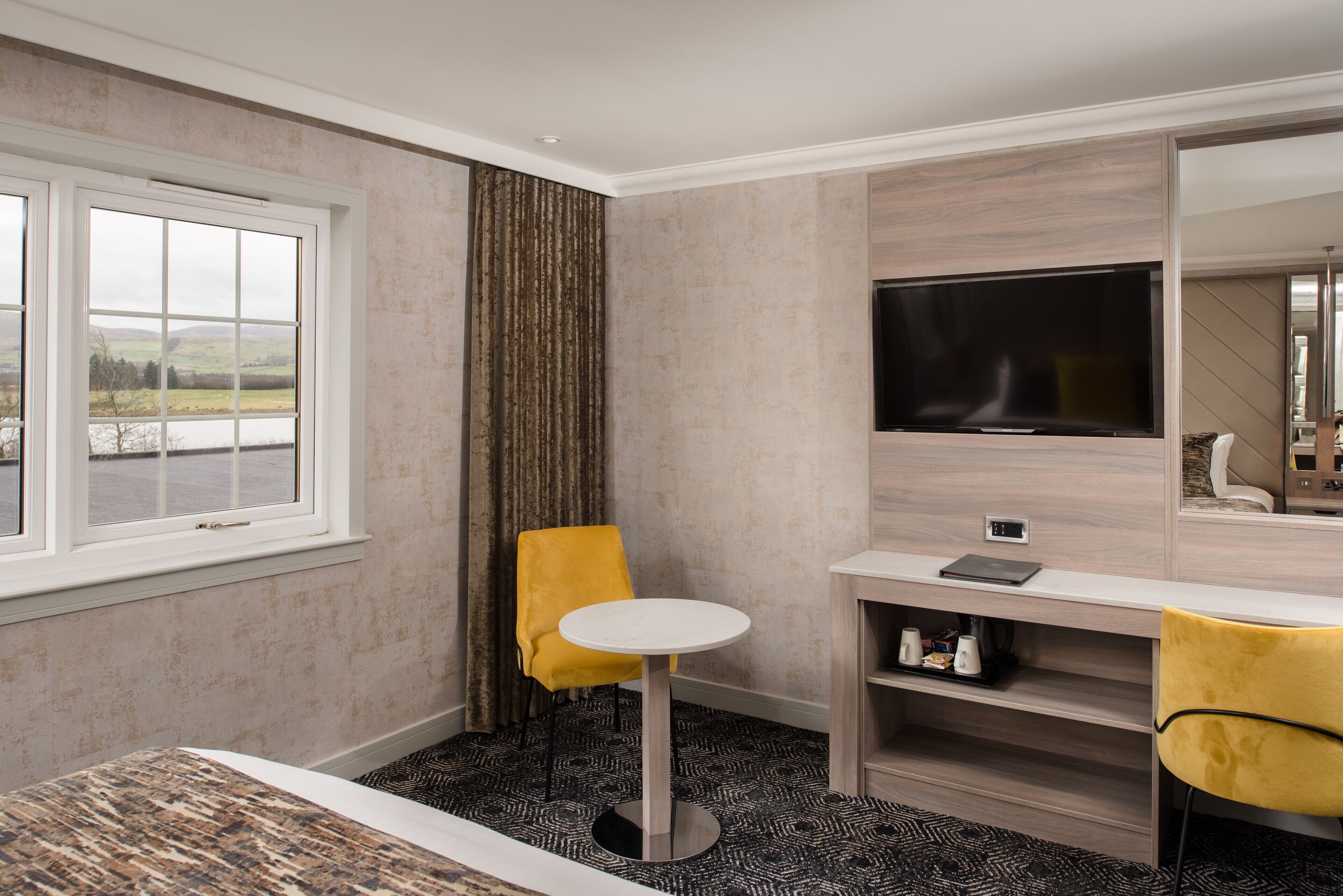 A hotel room with a large window, a round table, a yellow chair, a flat-screen TV, and a mirror.