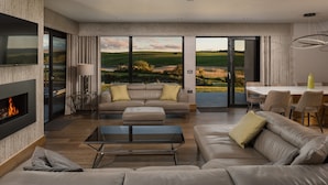 A modern living room with a fireplace, leather sofas, a glass coffee table, and a dining area with a view of the countryside.