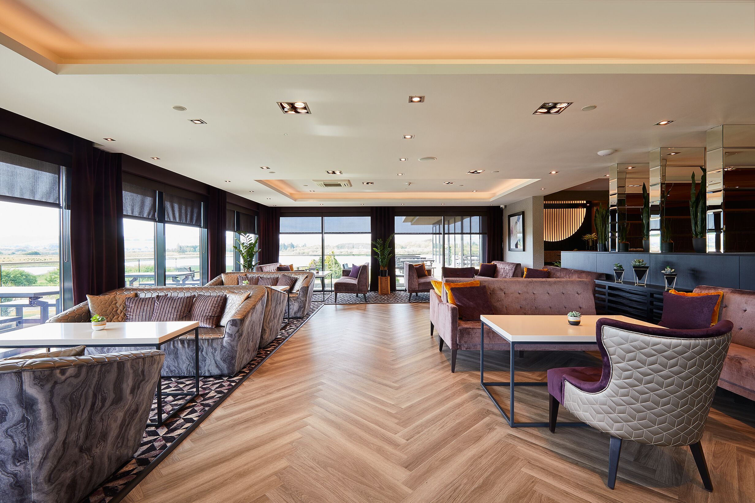 A spacious lounge area with seating arrangements, a herringbone wooden floor, and large windows offering a view.