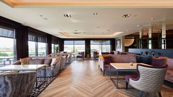 A spacious lounge area with seating arrangements, a herringbone wooden floor, and large windows offering a view.