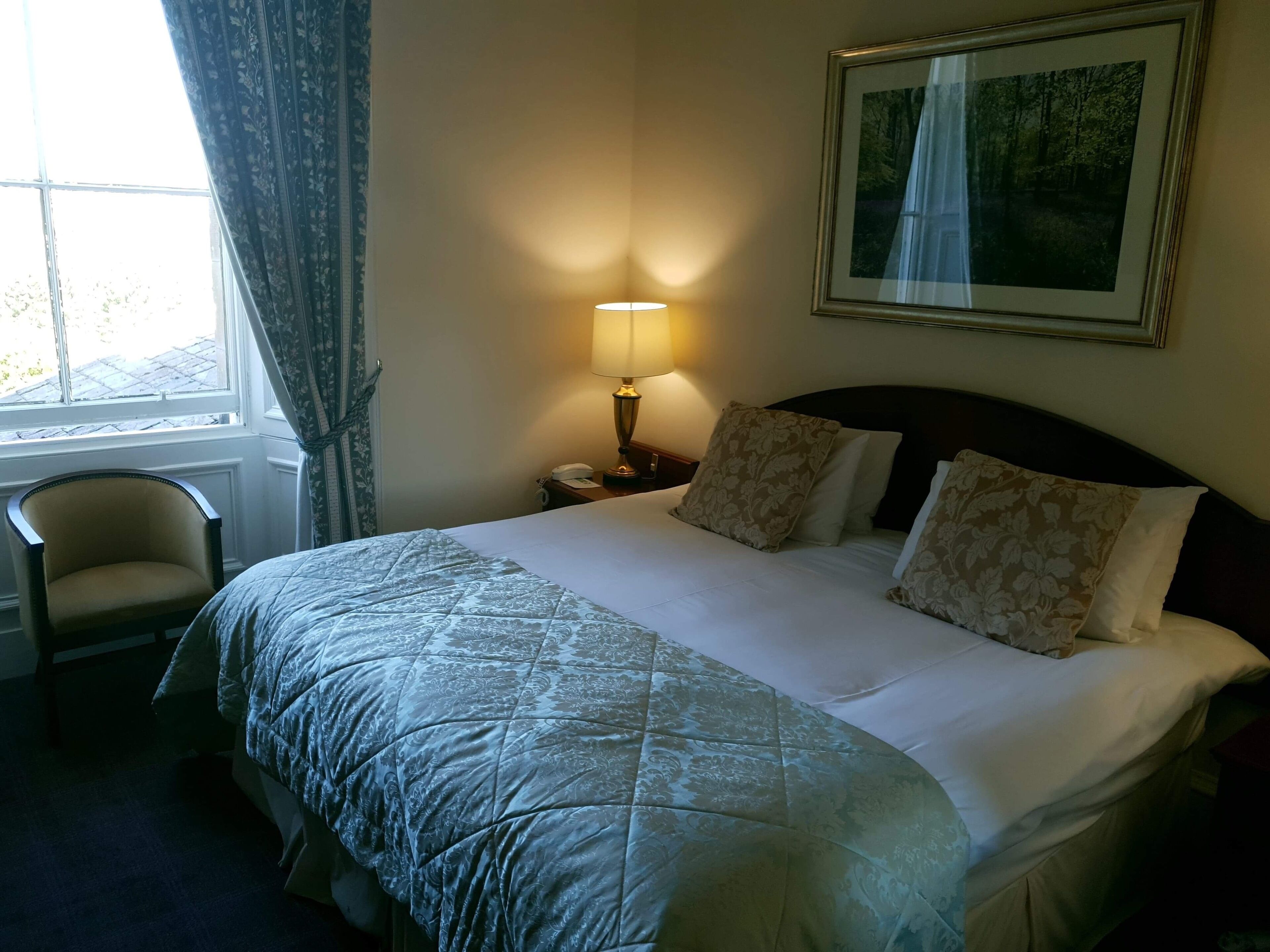 A hotel room with a large bed, a chair, a bedside table with a lamp, and a framed picture on the wall.