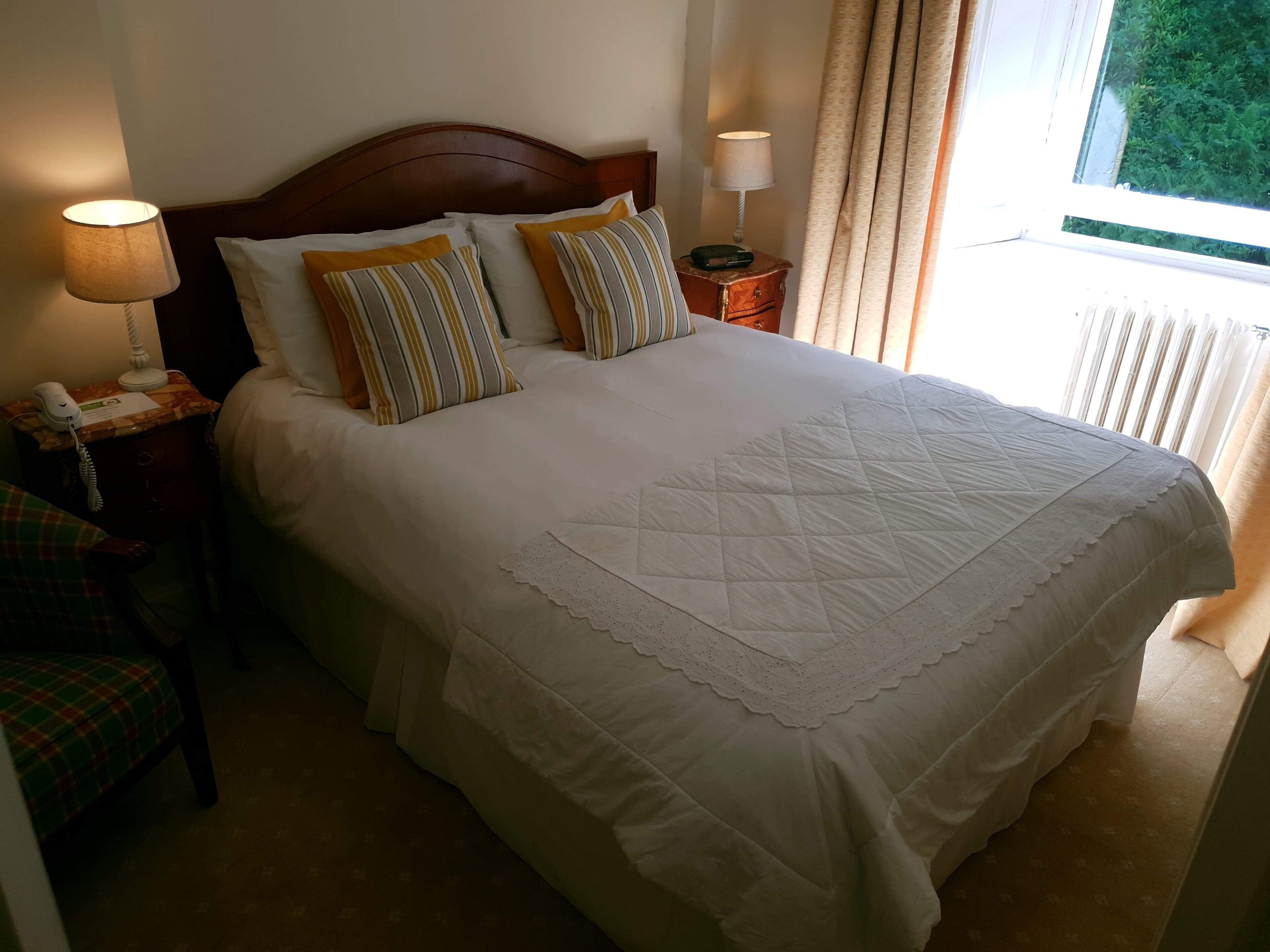 A neatly made bed with white linens and a quilt, a bedside lamp, a telephone, and a chair with a plaid cover.
