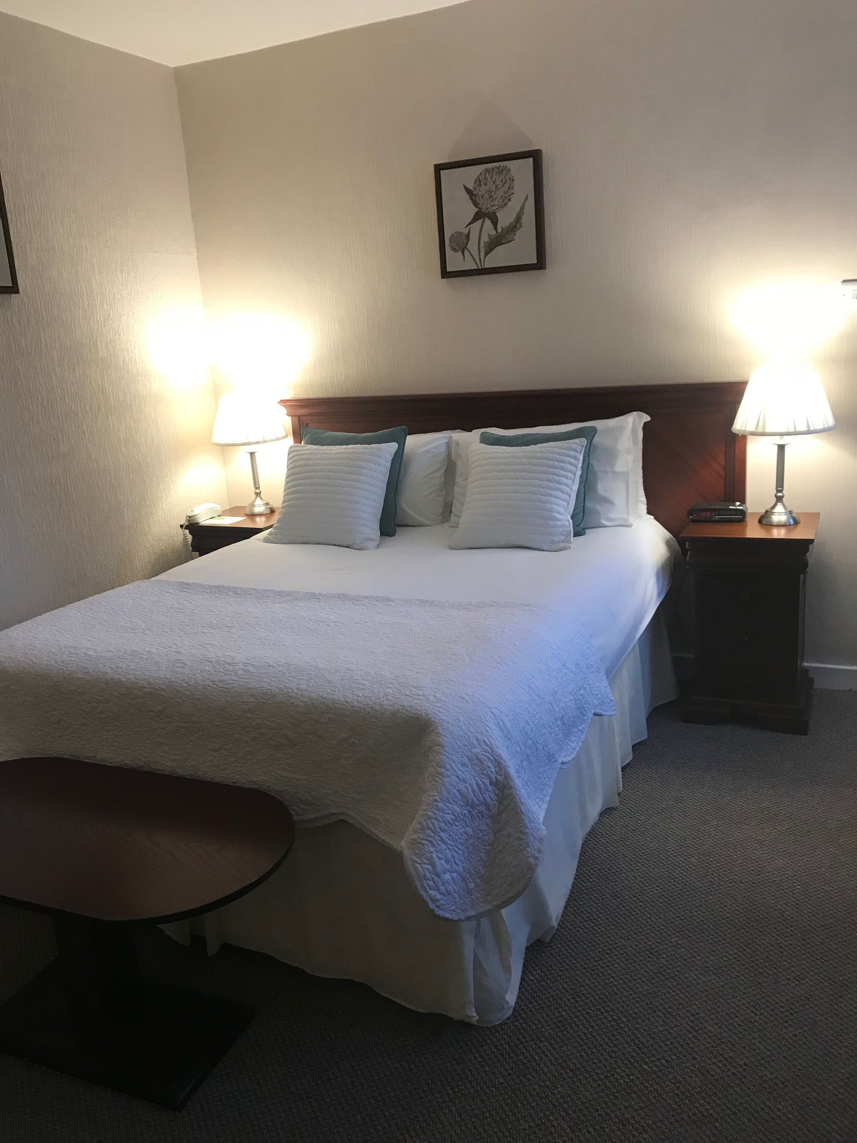 A neatly made bed with white linens and pillows, two bedside lamps, a small wooden nightstand, and a framed botanical print on the wall.