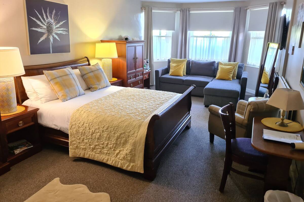 A hotel room with a bed, a sofa, a desk, and a chair. There is a lamp on the desk and a lamp on the nightstand.