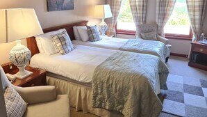 A hotel room with two beds, a chair, a lamp, a painting, and a window with curtains. - Mansfield House Hotel (Hawick)